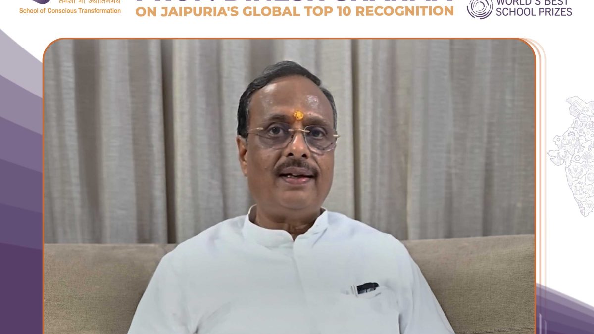 Video frame of a man in a white kurta with tilak, seated on a sofa, speaking, with a congratulatory banner and school logos above.