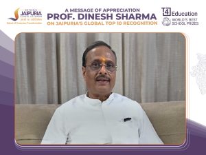 Video frame of a man in a white kurta with tilak, seated on a sofa, speaking, with a congratulatory banner and school logos above.