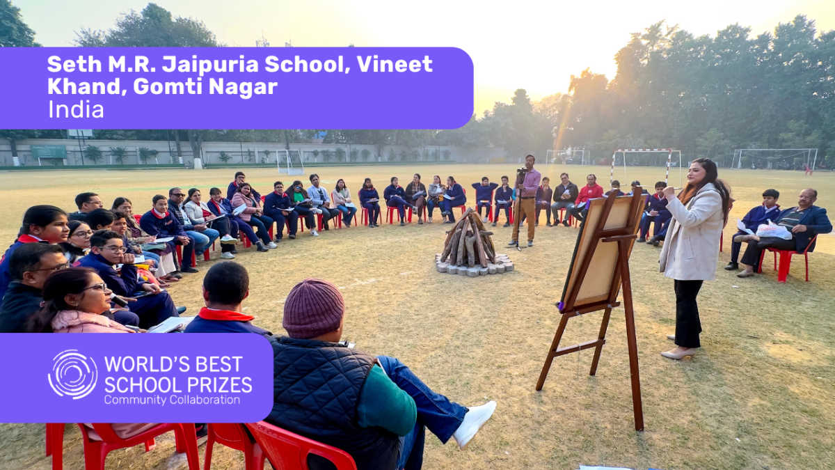 Students sit in a circle on red chairs in a school field, listening to a presenter by an easel, with banners displaying the school name and prize logos.