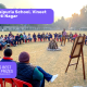 Students sit in a circle on red chairs in a school field, listening to a presenter by an easel, with banners displaying the school name and prize logos.