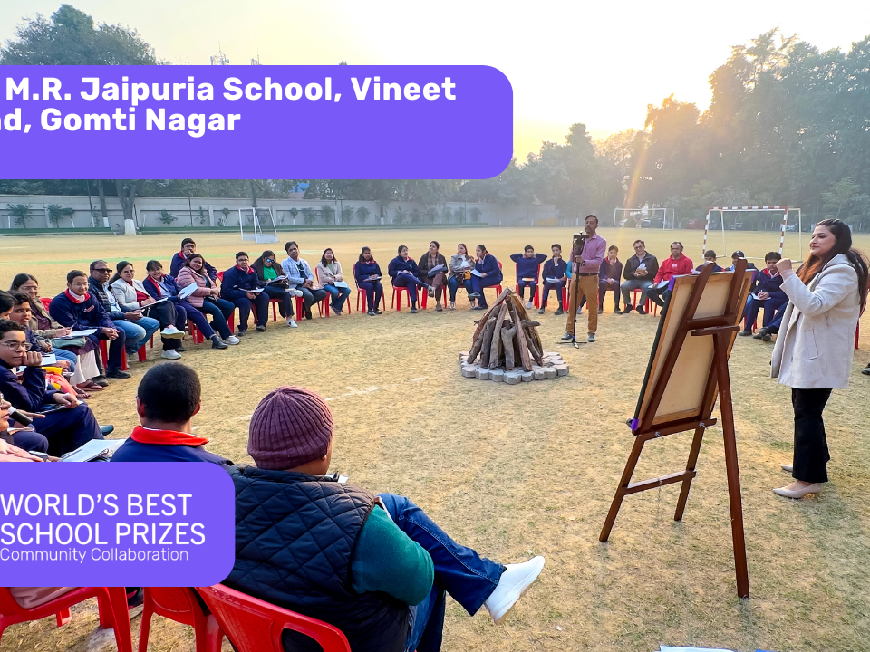 Students sit in a circle on red chairs in a school field, listening to a presenter by an easel, with banners displaying the school name and prize logos.