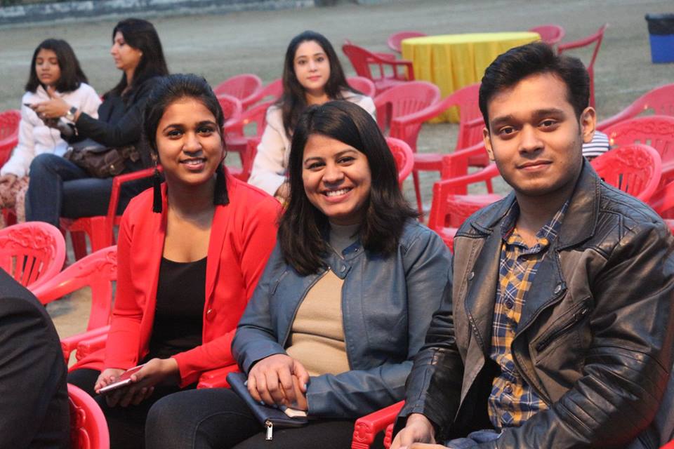 Three friends sit on red plastic chairs at an outdoor event, smiling at the camera: left woman in a red blazer, middle woman in a blue jacket, right man in a leather jacket.