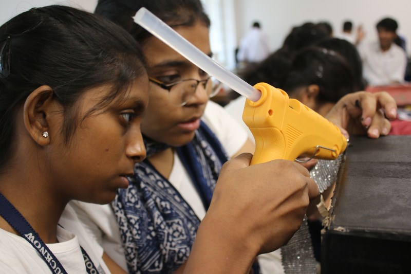 Two students in a classroom use a yellow hot glue gun to attach material to a craft project, concentrating on their work.