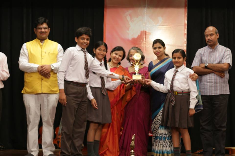 Group of students in school uniforms receiving a trophy on stage, flanked by teachers and parents.