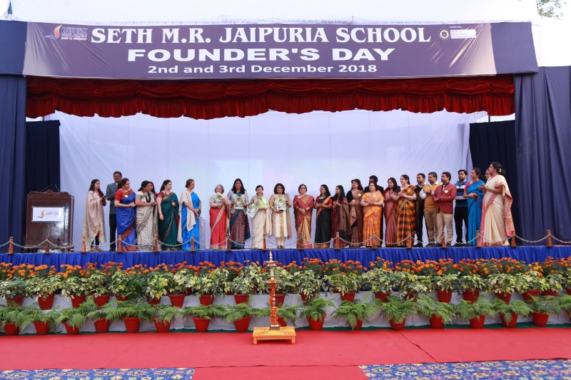 Group of people in colorful sarees and formal attire on a school stage during Founder’s Day ceremony, banner above reads 'Seth M.R. Jaipuria School Founder’s Day'