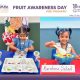 Two young girls participate in a fruit-themed classroom activity with labeled fruit cards at a Rainbow Salad station during Fruit Awareness Day.