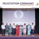 Group of award recipients and staff posing on stage after a felicitation ceremony, some holding certificates and a trophy