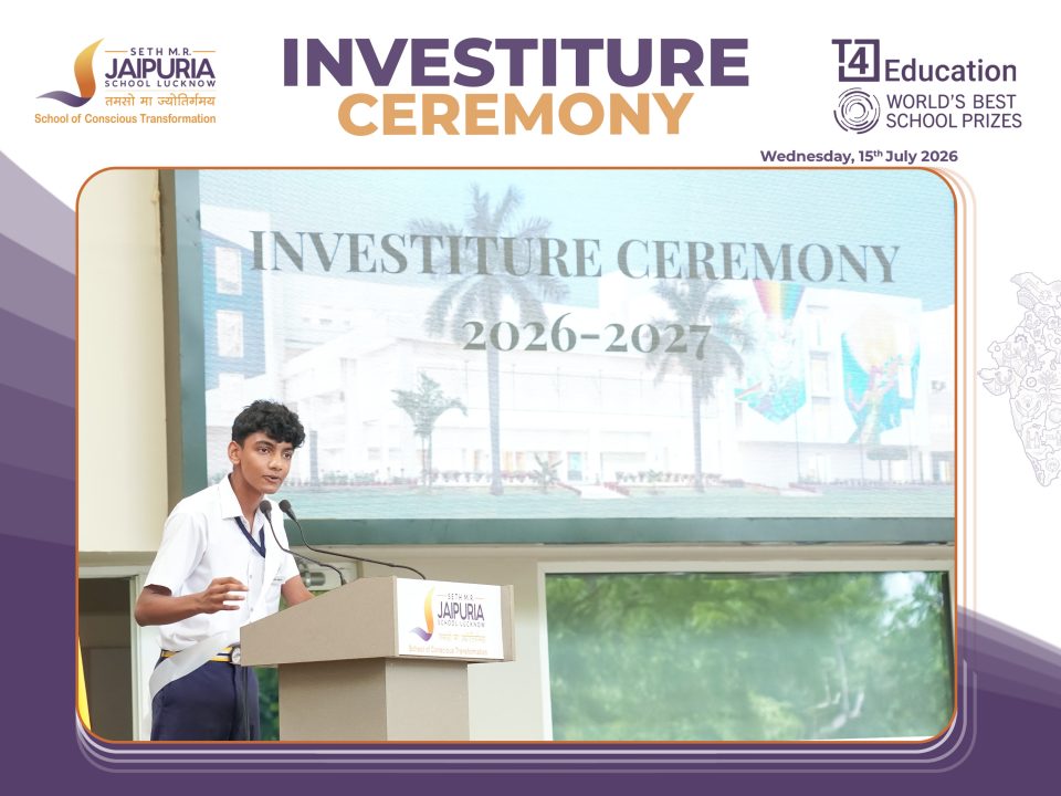 Boy in white shirt speaks at podium during school investiture ceremony; banner behind reads 'Investiture Ceremony 2026-2027' with school logos nearby.