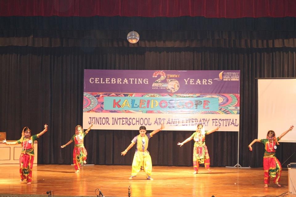 Five children in bright traditional costumes perform a dance on a stage, arms extended, in front of a banner that says 'Kaleidoscope' and 'Junior Interschool Art and Literary Festival' celebrating 25 years.