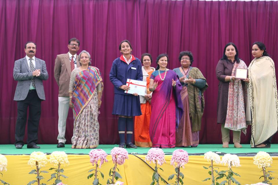 Group of nine people on a stage with a purple curtain; a student in a blue uniform holds a certificate while others clap and smile.