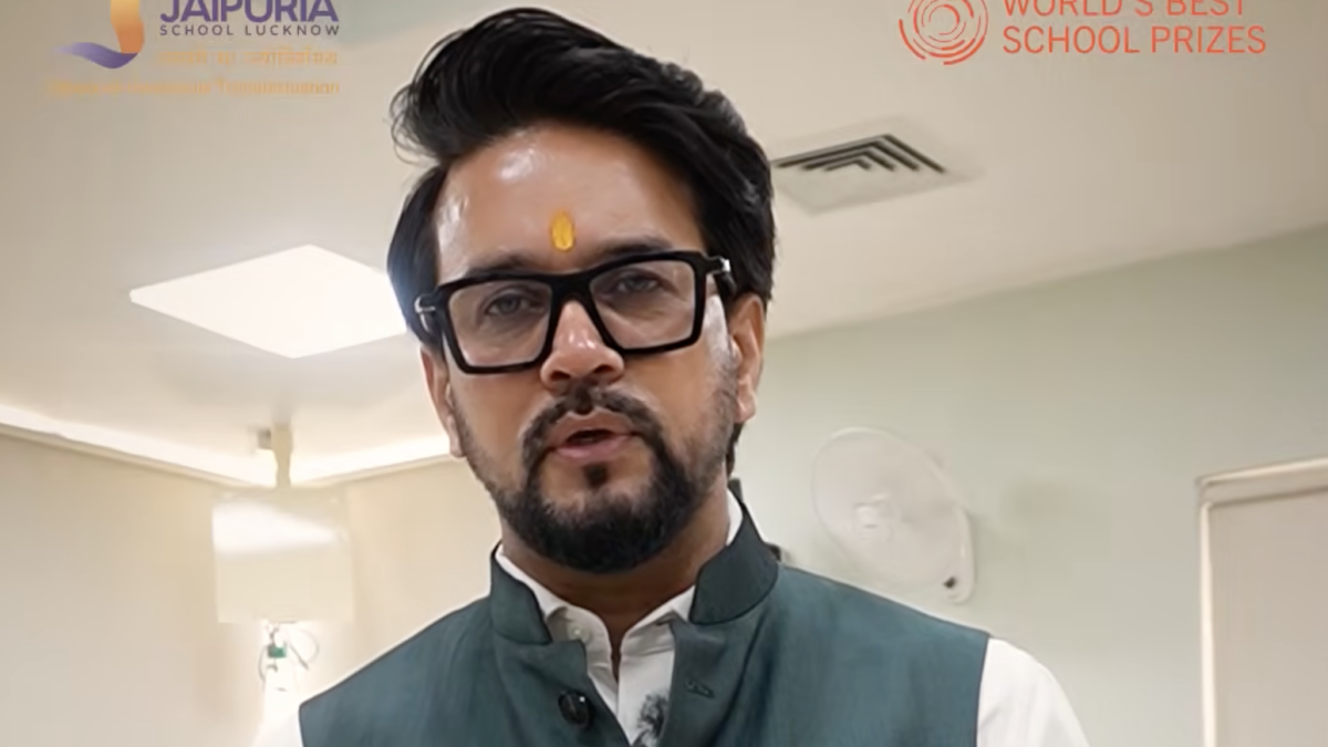 Man in a green vest and white shirt with a yellow tilak on his forehead, speaking in a hallway with school logos in the background.