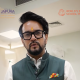 Man in a green vest and white shirt with a yellow tilak on his forehead, speaking in a hallway with school logos in the background.