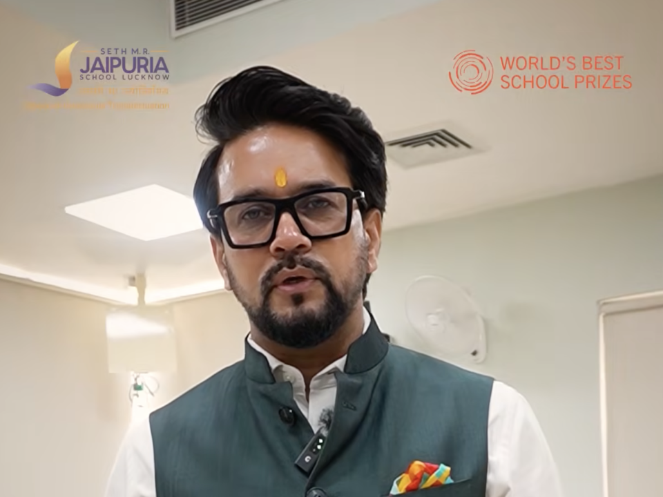 Man in a green vest and white shirt with a yellow tilak on his forehead, speaking in a hallway with school logos in the background.