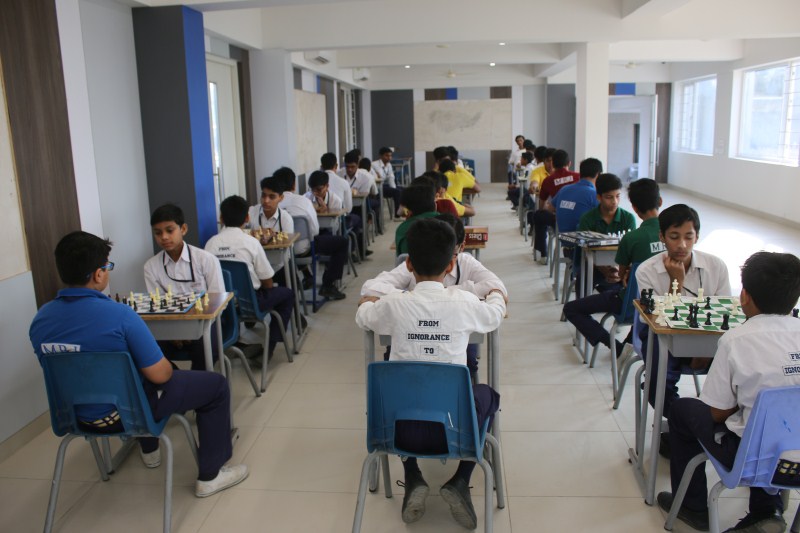 Students in a bright classroom seated in rows, playing chess in a school tournament paired across tables.