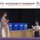 Woman speaks into a microphone on stage beside blue labeled recycling bins at a sustainability workshop.