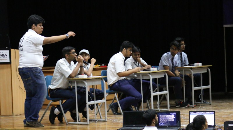 Instructor stands at left, gesturing toward students in white shirts seated at desks on a stage with laptops nearby.
