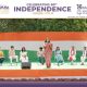 Group of students seated on a green stage as a woman in a saree speaks during a school Independence Day program.