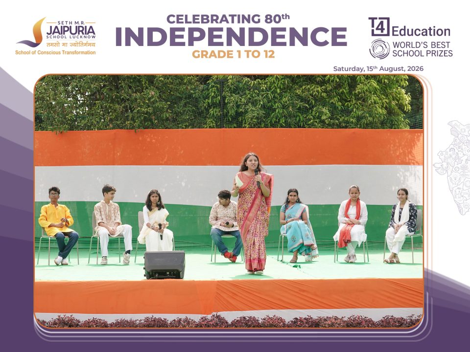 Group of students seated on a green stage as a woman in a saree speaks during a school Independence Day program.