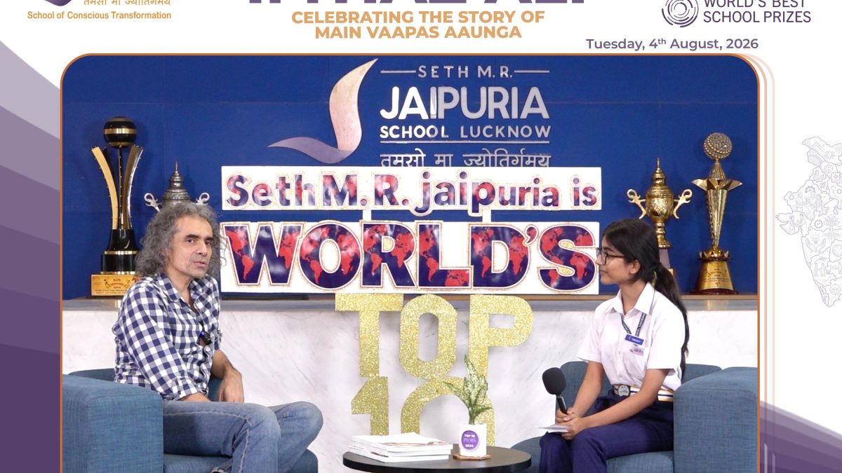 Two people sit in armchairs on a stage during a school event interview, with a blue backdrop and large trophies behind them, and a banner reading 'Seth M.R. Jaipuria School Lucknow' visible above.