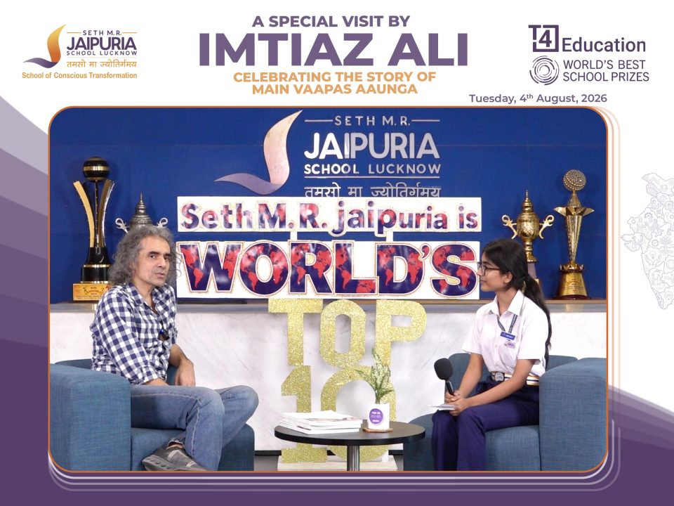 Two people sit in armchairs on a stage during a school event interview, with a blue backdrop and large trophies behind them, and a banner reading 'Seth M.R. Jaipuria School Lucknow' visible above.