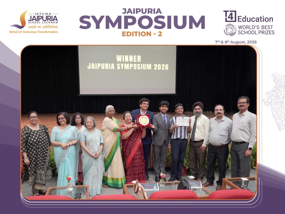Group of award recipients and attendees posing on stage at Jaipuria Symposium 2026 winner ceremony on screen behind them.