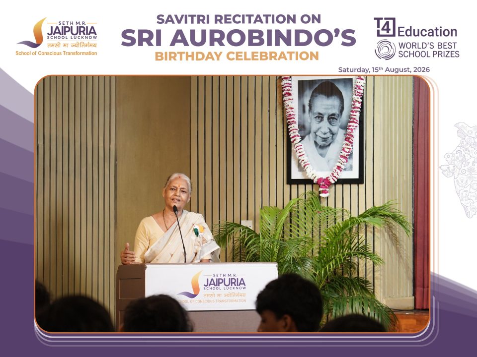Woman speaking at a podium during a celebration event, with a banner reading 'Seth M.R. Jaipuria School Lucknow' in the background.