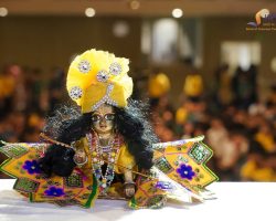 Colorful puppet with a yellow jeweled headdress and ornate costume on a stage; audience blurred in the background.