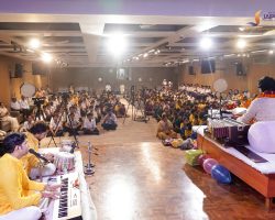 School concert in a hall with musicians at front and a large audience of students seated on the floor and chairs