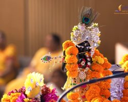 Ornate decorated Hindu deity idol adorned with peacock feather, jeweled crown, and orange marigold garlands on a ceremonial float; blurred festival crowd in the background