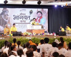 Speaker at a podium on stage during a cultural program, with musicians in yellow on the right and a large decorative backdrop behind him.