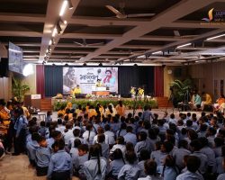 Auditorium filled with uniformed students facing a stage backdrop with a banner and speakers at the podium