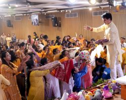 Man in cream traditional attire distributes gifts to a cheering crowd at a lively indoor celebration; attendees mostly wear yellow." ,