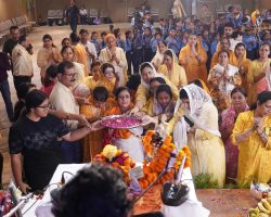 Crowd of people in yellow and white traditional outfits performing a ritual with a flower-filled platter, as children in blue uniforms watch in the background of a large hall.