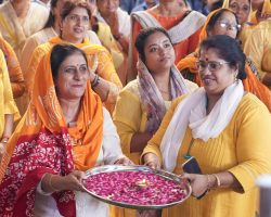 Women in yellow and orange attire carry a large silver tray of pink flower petals and a small lit lamp during a ceremonial gathering.