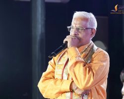 Speaker in orange traditional attire with glasses, holding a microphone on stage; logo in the top-right corner.
