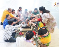 People sit in a circle around a small grill, cooking and sharing food outdoors with bowls of ingredients nearby.
