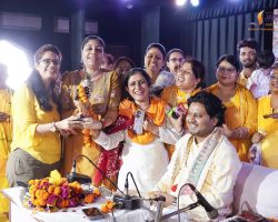 Group of people in yellow traditional outfits posing and smiling on a stage with flowers and garlands.