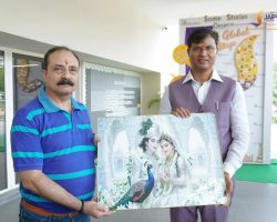 Two men stand outdoors holding a framed painting of Radha and Krishna with a peacock, in front of a school backdrop with a Jaipuria School Lucknow sign visible behind them.