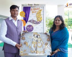 Two people in formal attire stand outdoors, presenting a large religious painting of Hindu deities while smiling at the camera.