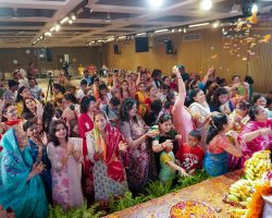 Crowd of people in colorful Indian attire celebrating with petals and flowers at a festival indoors.