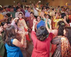 A large group of people in colorful Indian clothing dancing and celebrating indoors, with confetti on the floor and smiles on many faces.