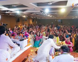 Crowded hall during a festive ceremony with people in colorful traditional attire around a long, garlanded table with offerings at the front.