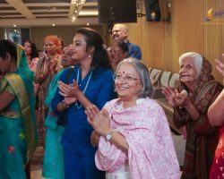 Group of Indian women and a man clapping at an indoor event, colorful traditional attire and smiles evident in a hall with a logo in the corner.