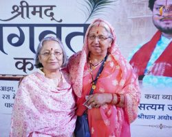 Two elderly Indian women in pink traditional outfits pose together on a stage with a Hindi banner in the background, smiling at the camera.