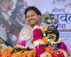 Smiling man in beige clothing at a festival, surrounded by vibrant garlands and a decorated idol in the foreground.