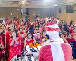 Crowd of women in colorful sarees at an indoor cultural event; cameras on tripods and a presenter seen from behind in the foreground.