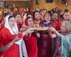 Women in colorful saris gather for a flower-ritual ceremony, passing a tray of petals together in a crowded indoor hall.