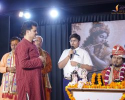 Man in a white shirt speaks into a microphone on a decorated stage, flanked by others in traditional attire, with orange marigold garlands and a large backdrop image of a goddess.