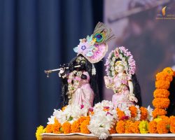 Ornate Hindu idols (Krishna and Radha) dressed in pink with floral headpieces, Krishna playing a flute, surrounded by orange marigold garlands on a stage.