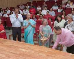 People in a hall participate in a traditional lamp-lighting ceremony at a wooden table, with an audience seated in red chairs behind them.